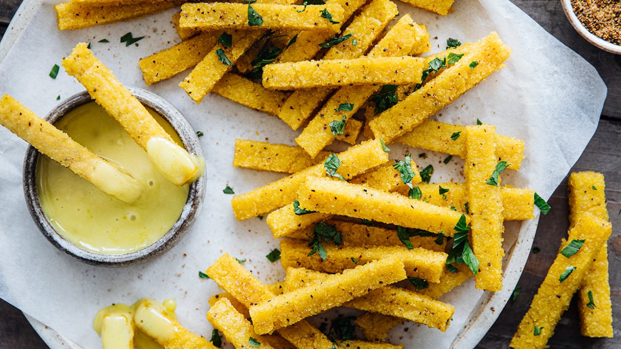 Sticks aus Polenta sind auf einem Teller angeordnet mit einen Dipp in einer kleinen Schale. 